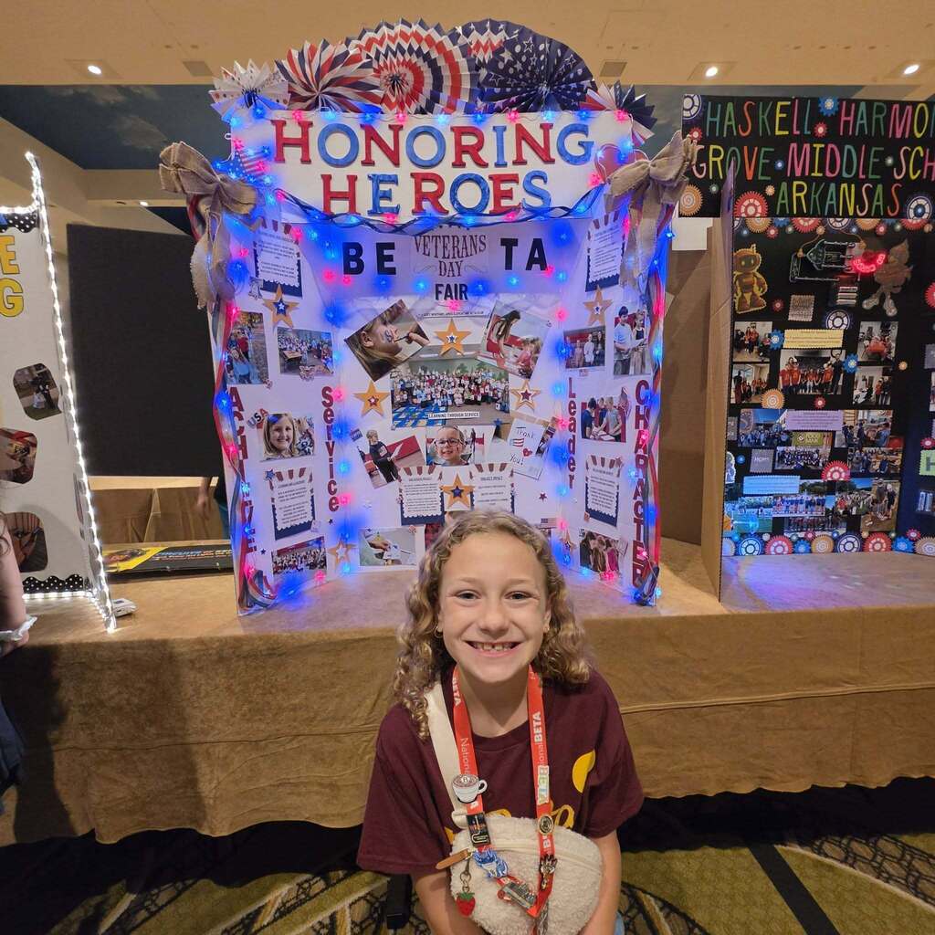 A smiling child stands before a large display titled "Honoring Heroes," adorned with photos and blue lights.