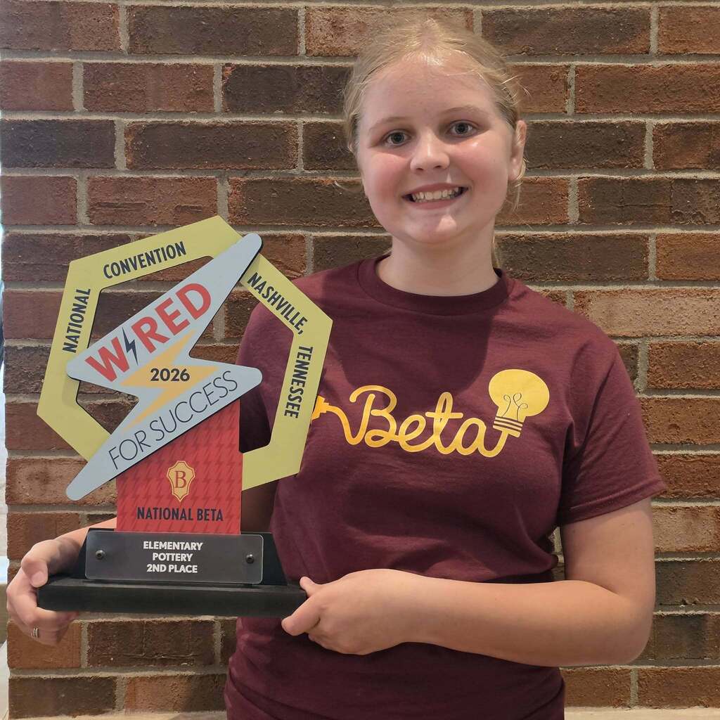 A young person holds a trophy in front of a brick wall. The trophy reads "Wired for Success" and "National Beta Convention."
