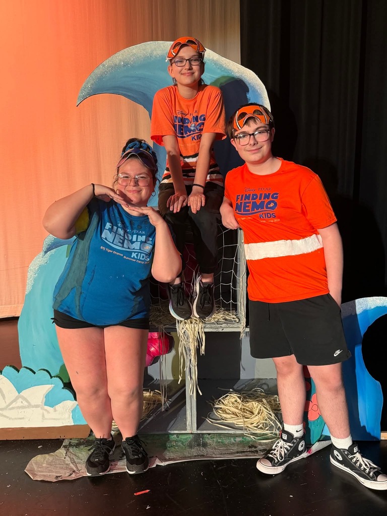 Three individuals on a stage, two wearing orange shirts and one in blue, posing with a blue and orange wave-like backdrop.