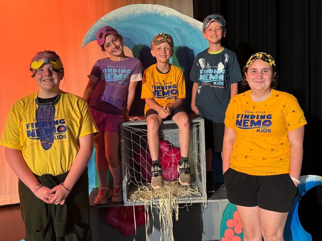 Six children wearing yellow t-shirts stand behind a metal box with a net on top. They appear to be on a stage.