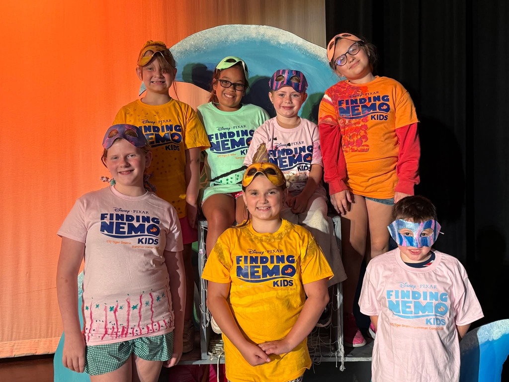 A group of children wearing themed shirts and head coverings stand together on a stage.
