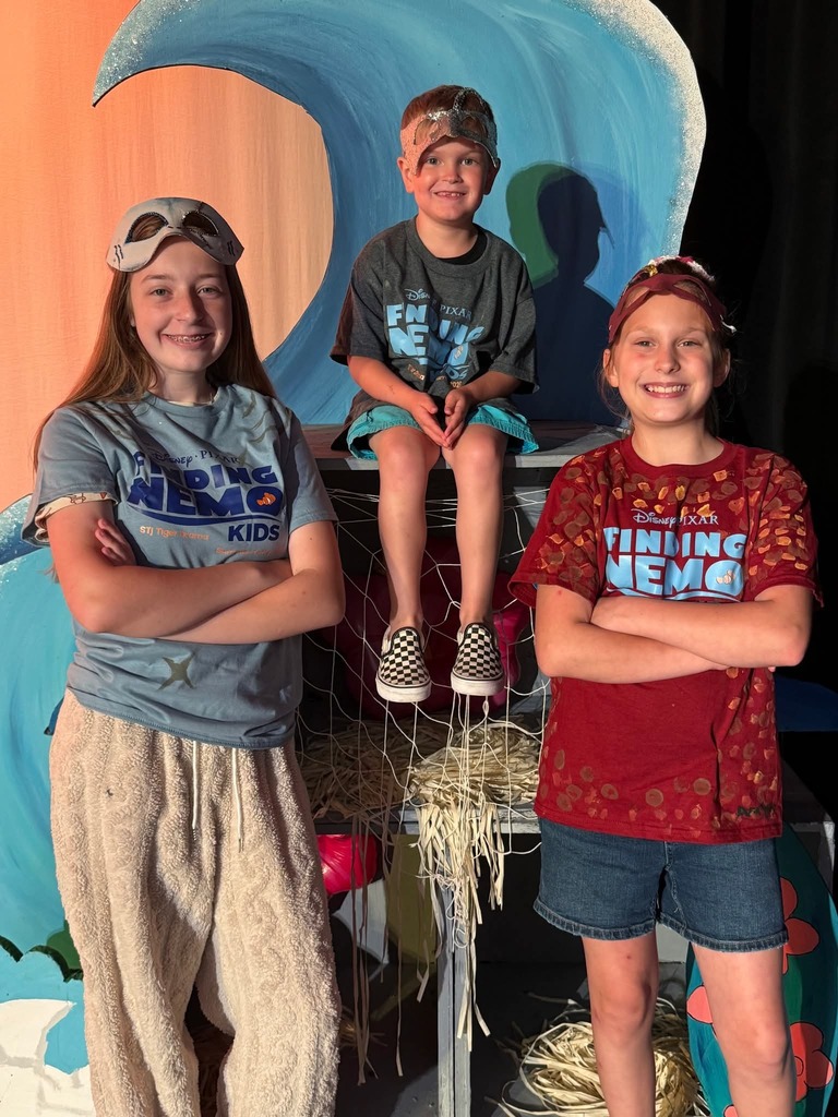 Three kids in costume stand on a stage, smiling. The girl on the left wears goggles and a gray shirt.