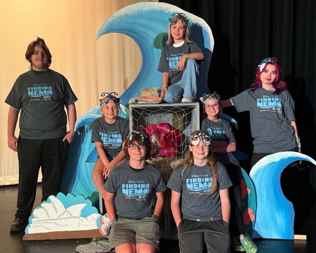 A group of people in matching t-shirts pose on a stage, with a wave backdrop and props.