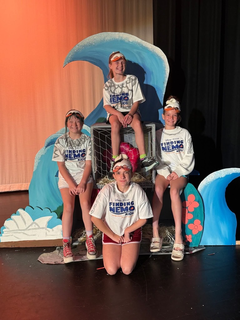 Four individuals in white shirts pose for a photo on a stage with a blue wave backdrop.