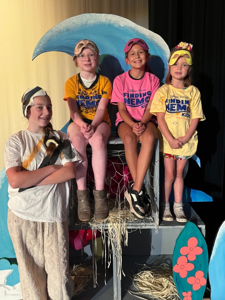 Five children stand on a stage with a wave backdrop. Two are seated, one holding a prop. The others stand with arms crossed and legs apart.