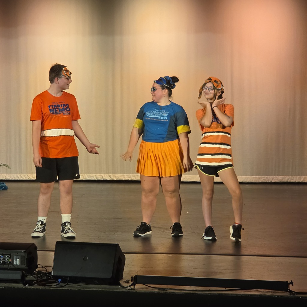 Three people on a stage. The person on the left wears an orange shirt. The middle person wears a blue shirt. The person on the right wears a striped shirt.