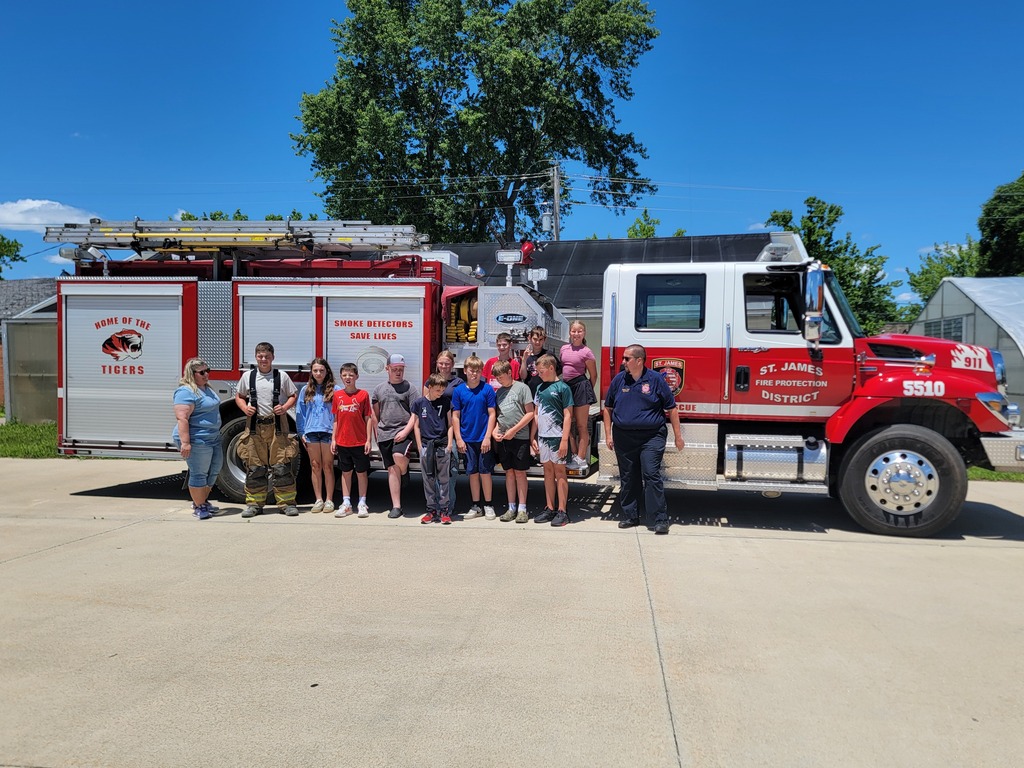 People stand in front of a firetruck with trees and a building in the background.