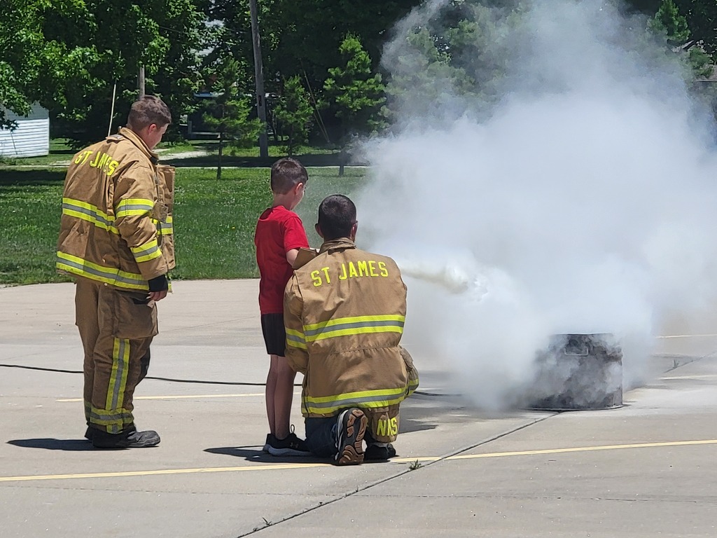 Three firefighters in uniforms, one kneeling, one standing, and a child nearby. Smoke rises from a black object on the road.