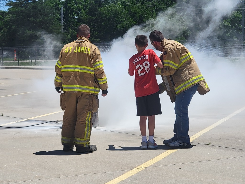 Three firefighters with a child in front of them. A boy in a red shirt stands between two firefighters.