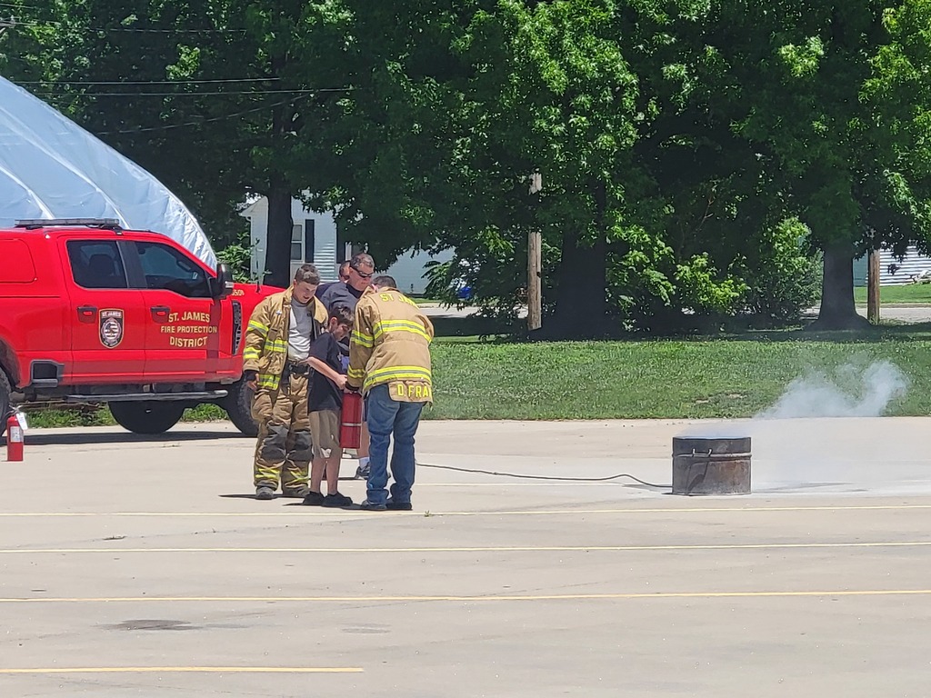 Three people in safety gear near a red truck. One person stands behind a barrel emitting smoke. Background includes trees and grass.