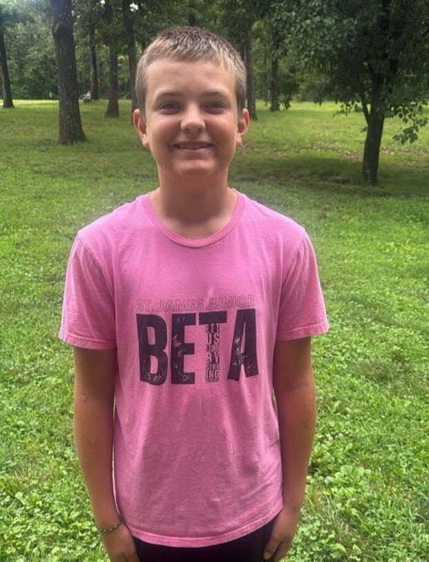 A boy in a pink T-shirt with black letters reading "BETA" stands in a grassy field with trees.