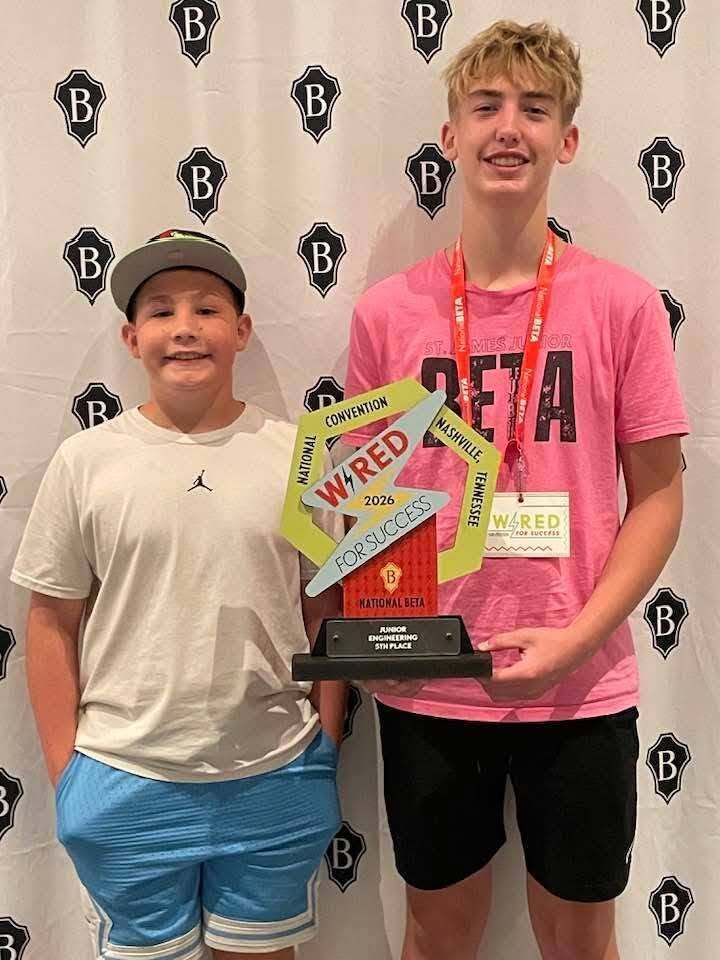 Two young boys stand next to each other holding a trophy. One wears a white shirt, the other a pink shirt. Both wear caps and sneakers.