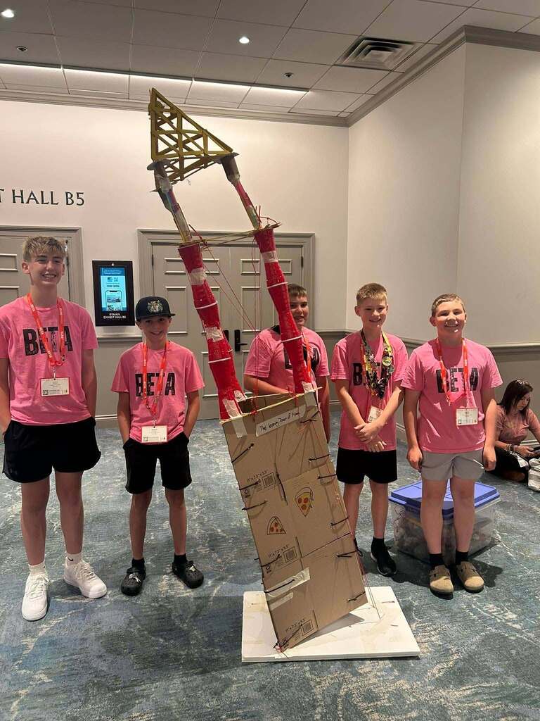 Five boys in pink shirts and black shorts pose next to a cardboard structure. The backdrop shows a hallway.