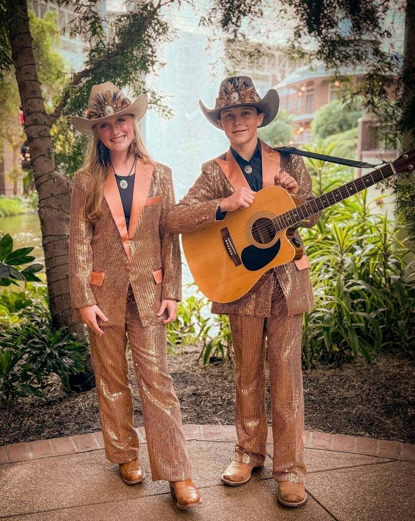 Two people in matching suits and hats standing on a patio, one holding a guitar.