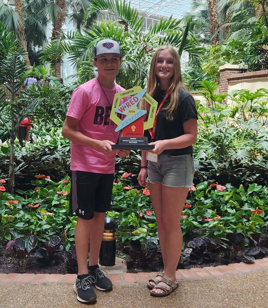 Two individuals pose outdoors, holding a trophy. A person wears a pink shirt and black shorts. Another person wears a black shirt and gray shorts.