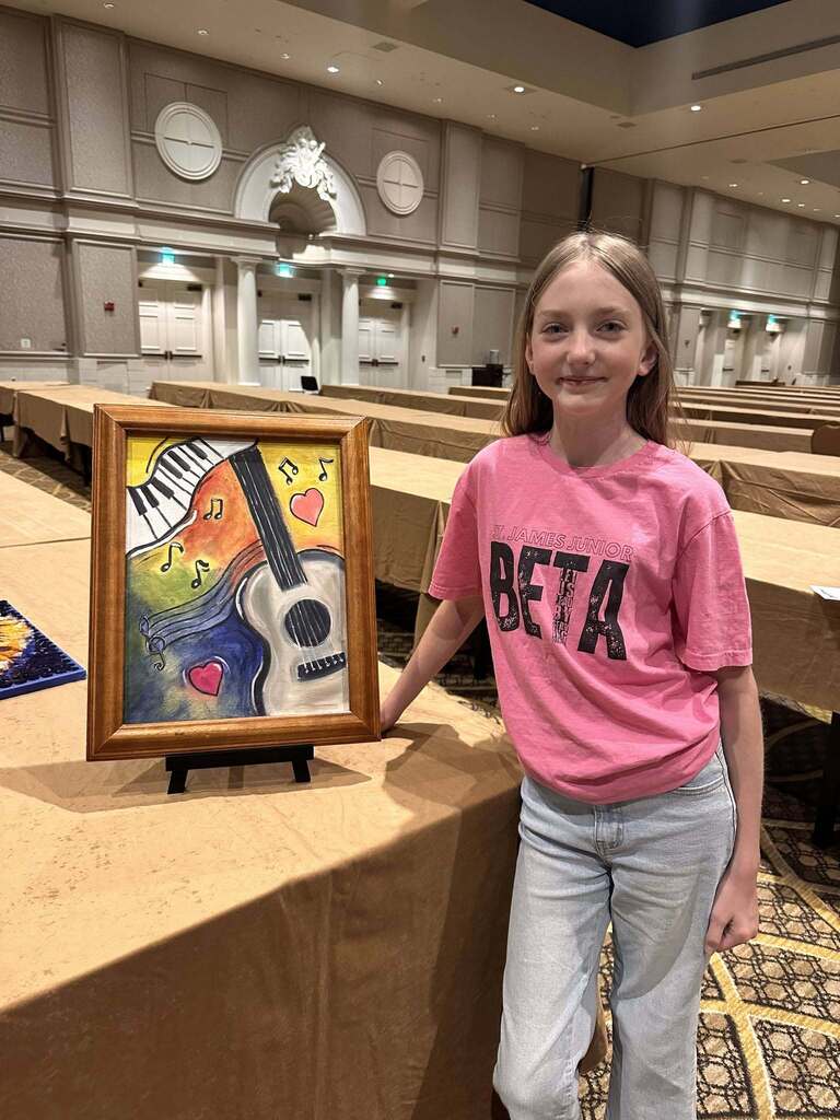 A girl stands next to a table with a framed painting of a guitar and piano, wearing a pink shirt.