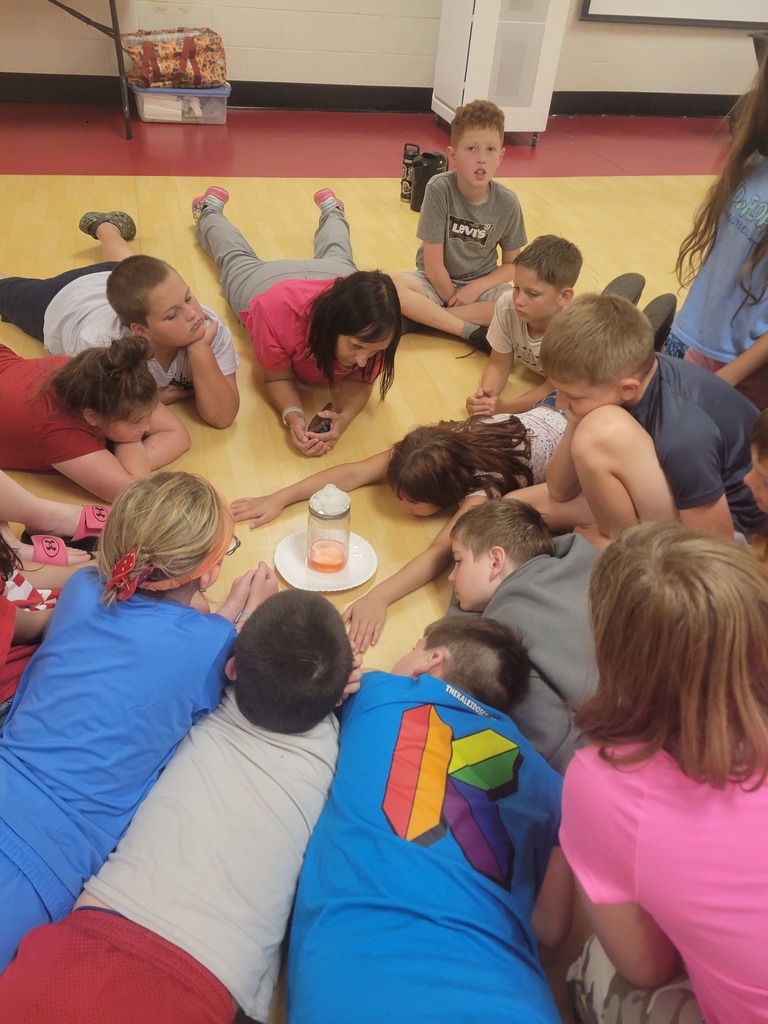 A group of children and adults sit on a floor in a circle. A woman holds a candle.