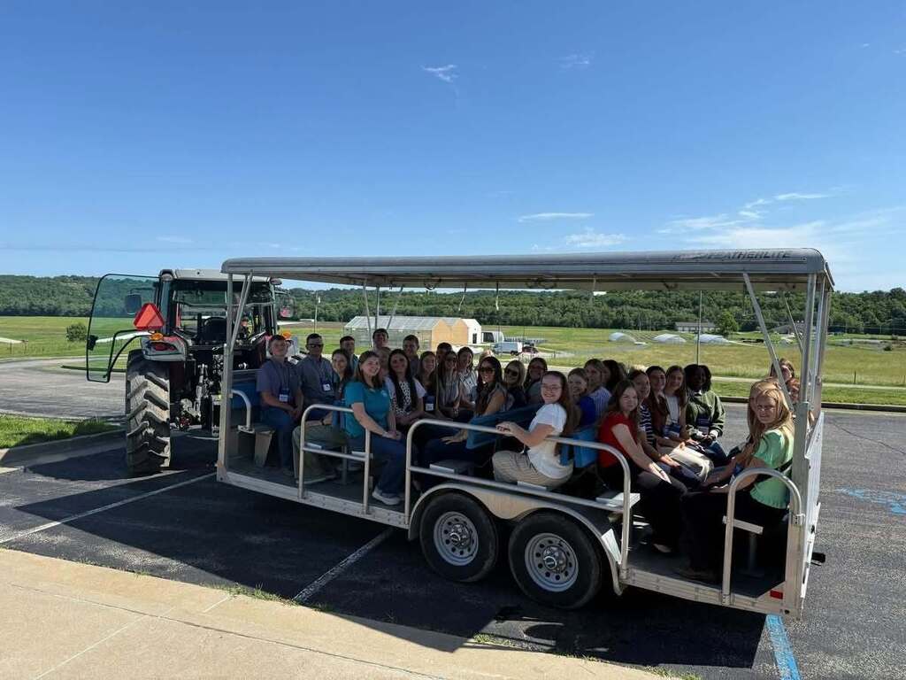 A group of people seated in a covered trailer being pulled by a tractor. Background shows a grassy area and distant trees.