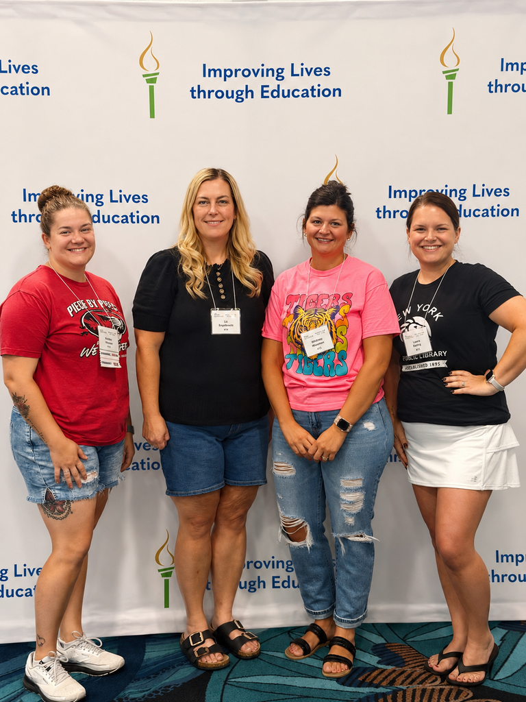Four women stand next to each other in front of a banner. Each wears a lanyard.