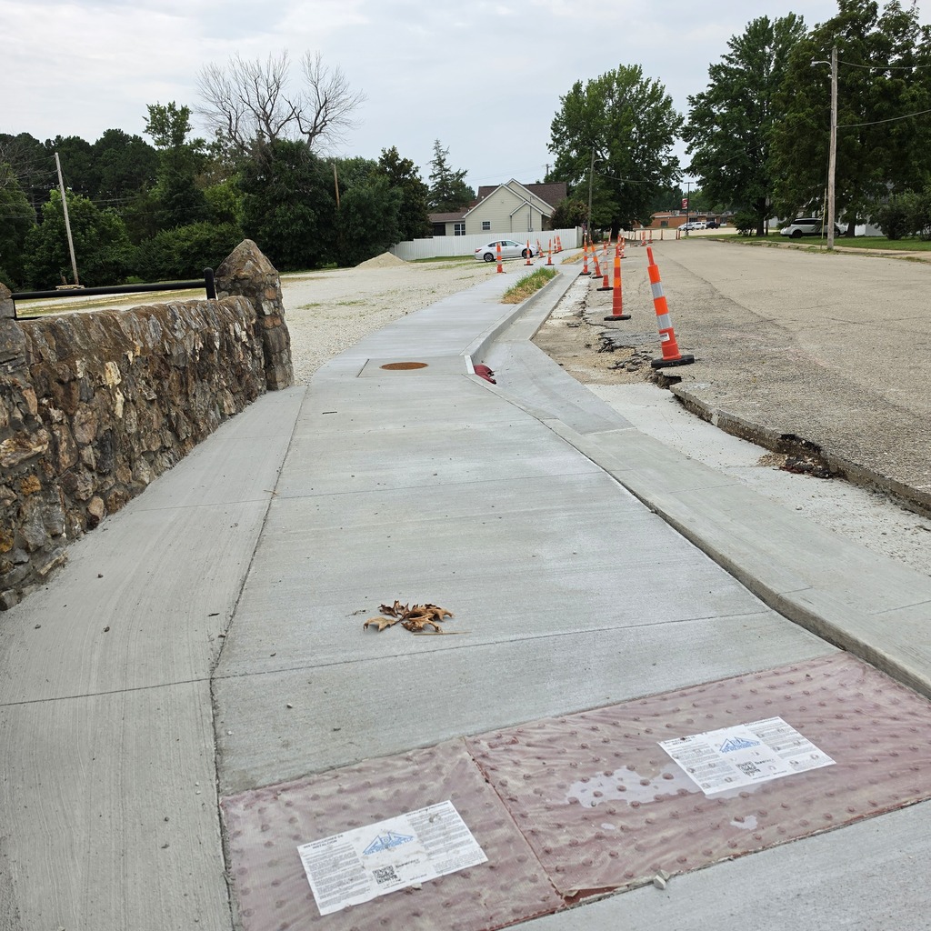 A concrete pathway with a stone wall on the left. Orange cones line the path, and there is a sign on the concrete.