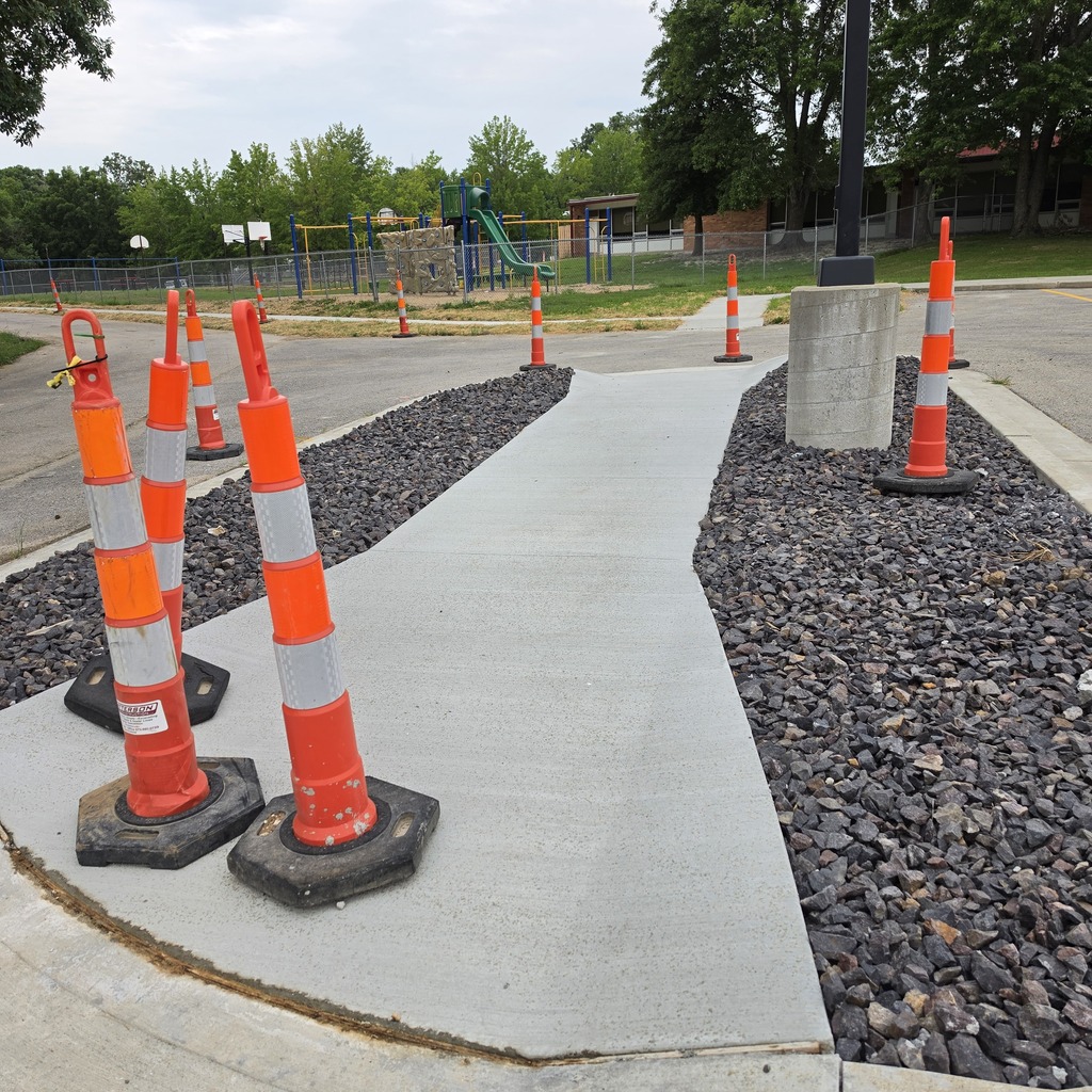 Sidewalk with orange and white cones on the side, gravel, and a playground in the background.