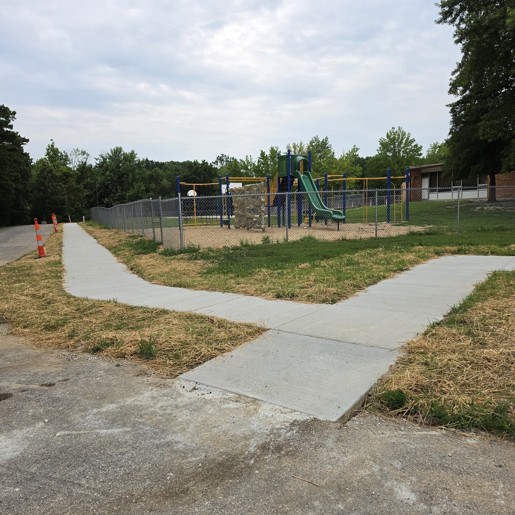 A playground with a slide, fence, and path. The area is surrounded by grass and trees.