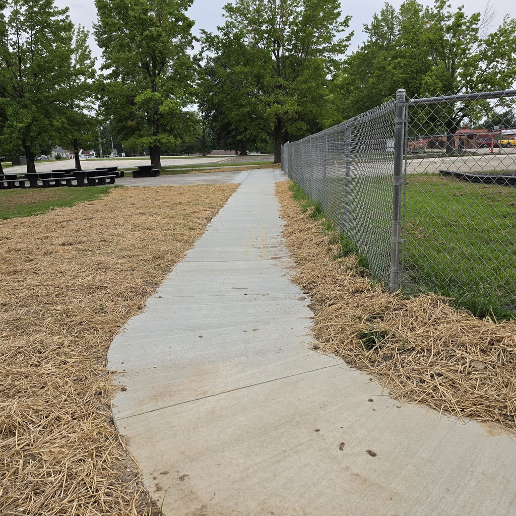 Concrete walkway with straw on the sides near a metal fence. Trees and grass are in the background.