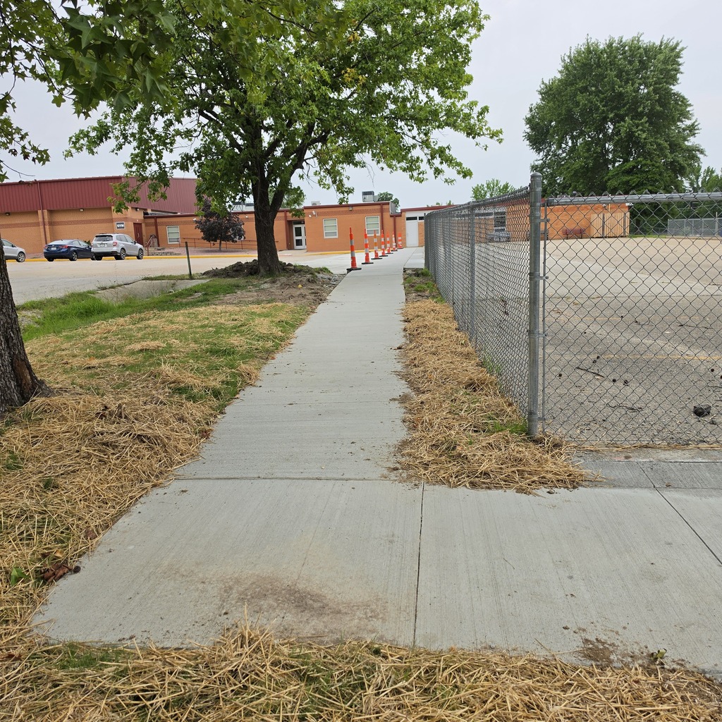 A walkway leads to a building with a fenced-off area behind it. Buildings, trees, and cars are in the background.