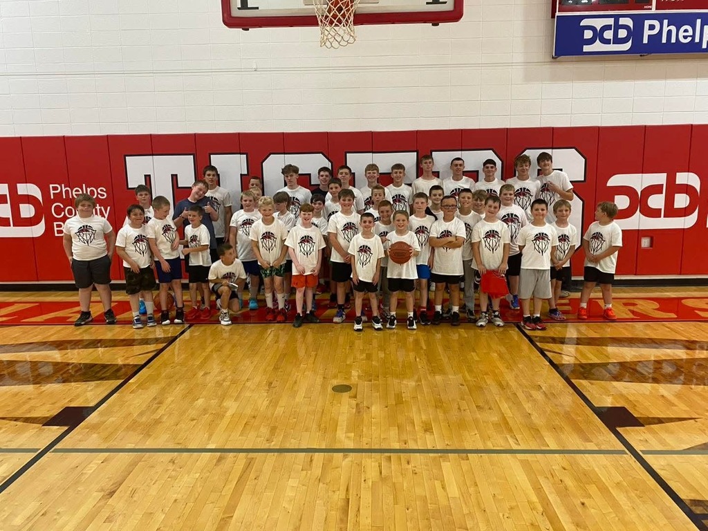Group of young basketball players in white T-shirts posing on a basketball court with red wall and black floor.