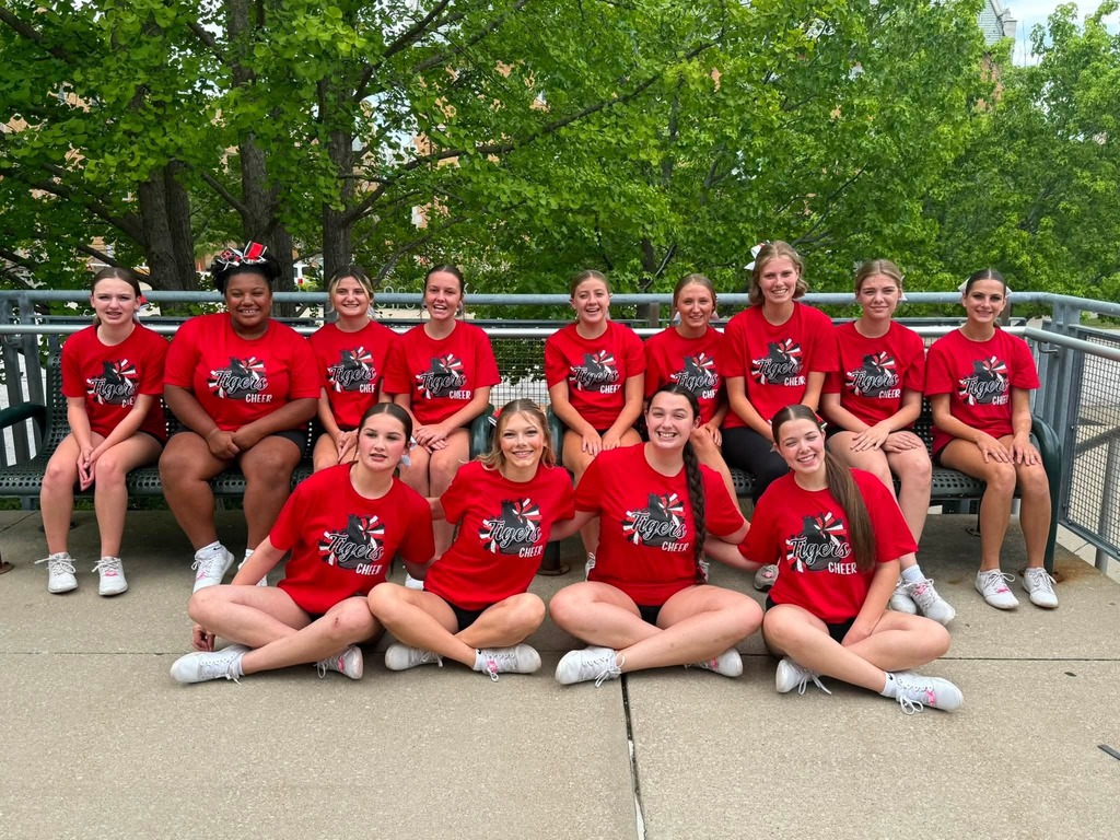 Group of young women in red t-shirts and shorts, sitting and kneeling, posing for a photo on concrete.