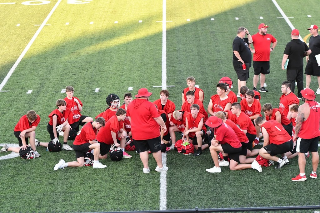 Group of players in red jerseys on a field, with coaches in red hats and black shirts nearby.