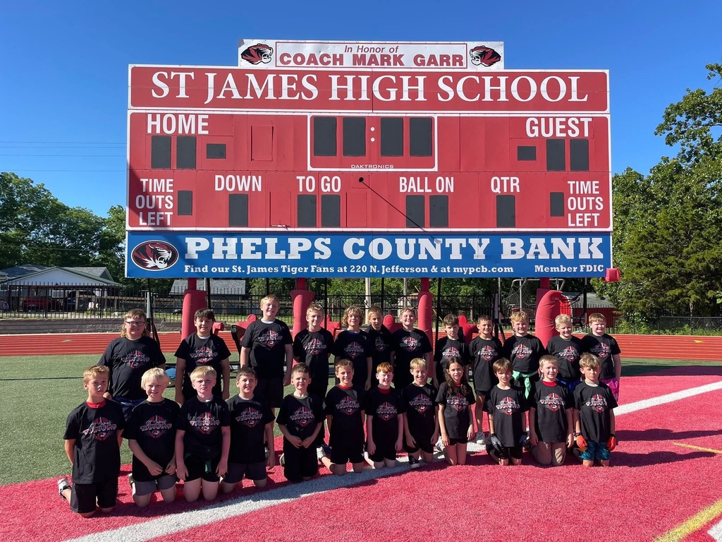 A team in black jerseys stands in front of a scoreboard for St James High School. A sponsor's name is displayed on the lower banner.