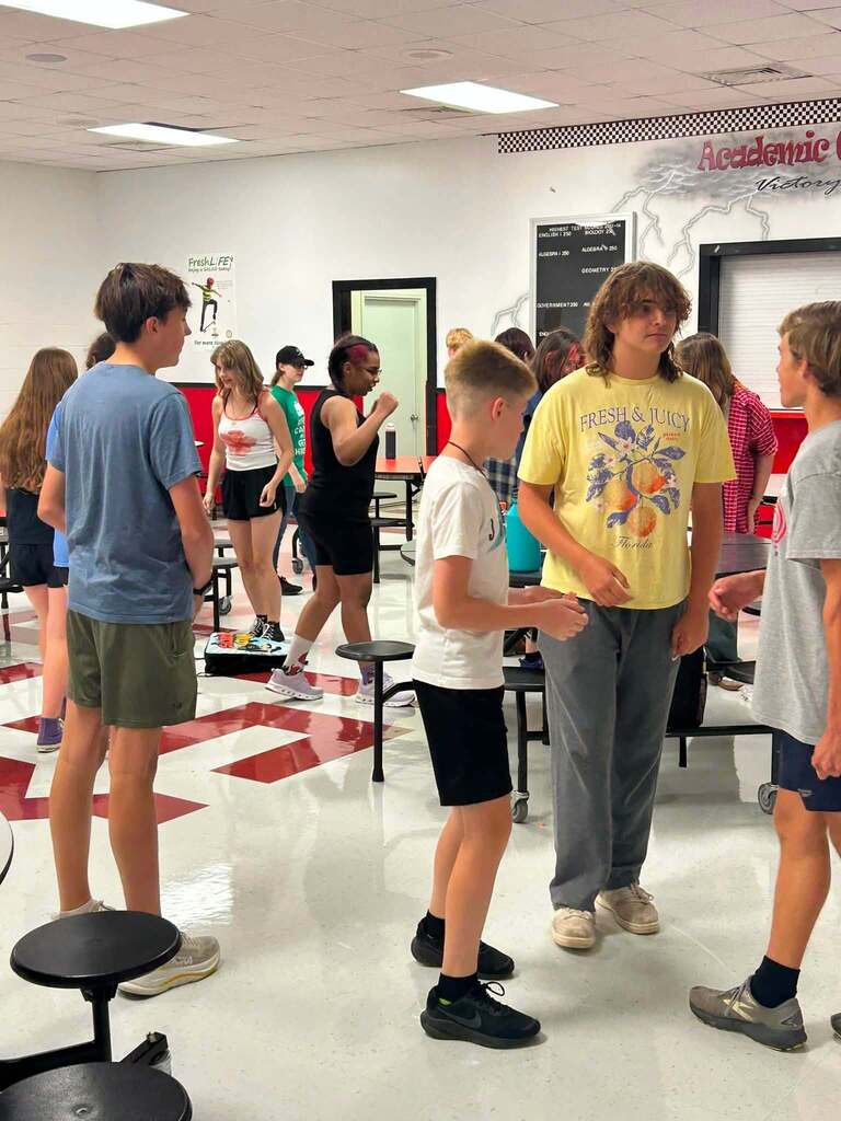 Group of students conversing in a school cafeteria. Tables and chairs are arranged. The floor has a red and white pattern.