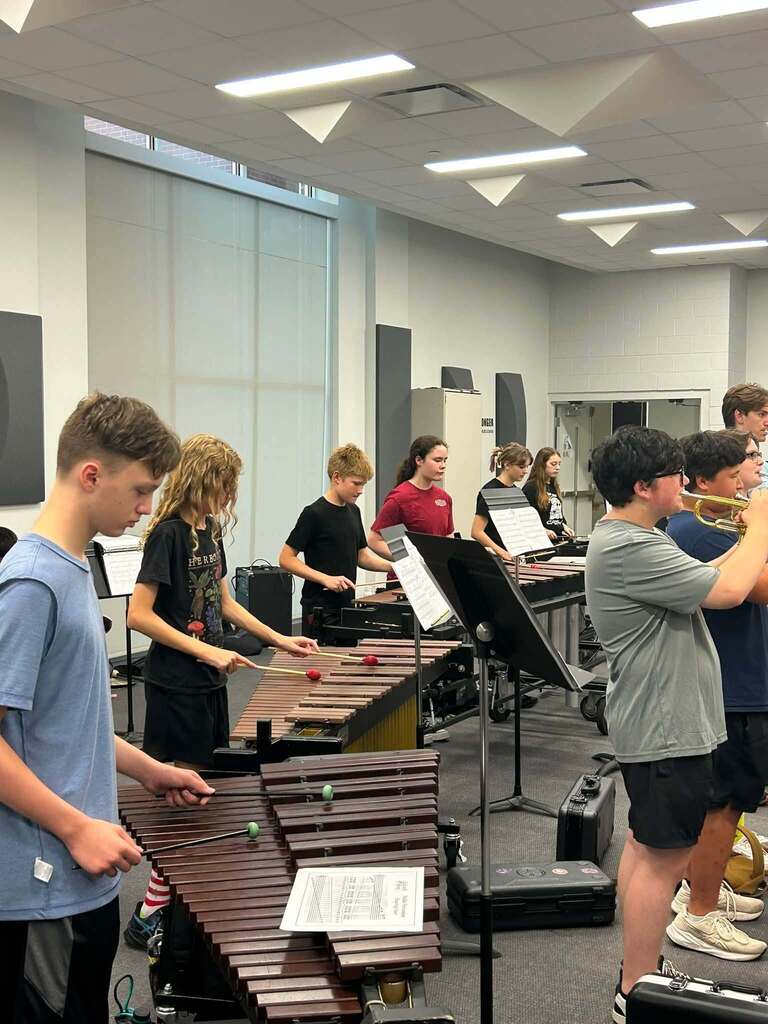 A classroom with music students. Some play trumpets and others play xylophones. A music stand with sheet music is in the foreground.