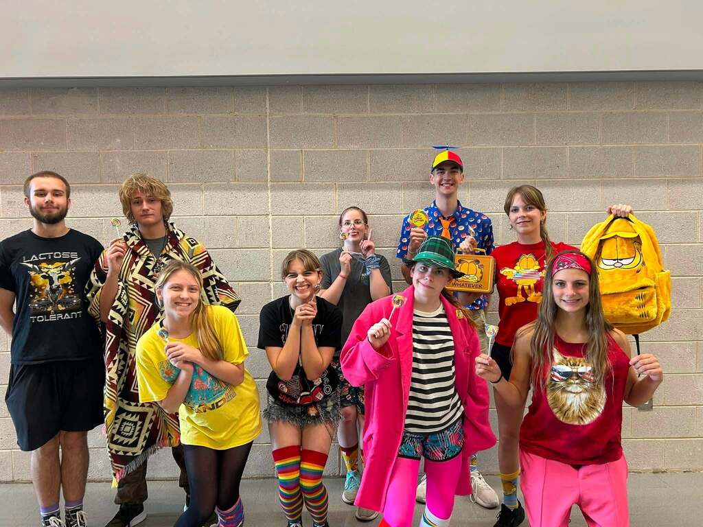 A group of people, some in colorful and quirky costumes, standing against a light-colored wall.