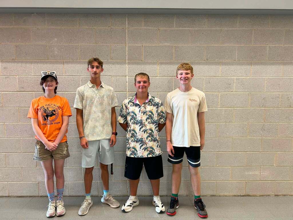 Five people stand in a row against a light gray wall, each wearing different colored shirts and shorts.