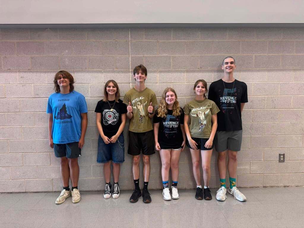A group of six young people stands in a row against a white wall, wearing casual clothing.