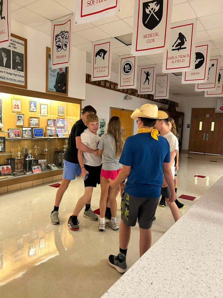 A group of students engage in a team-building activity in a school hallway with trophies displayed.