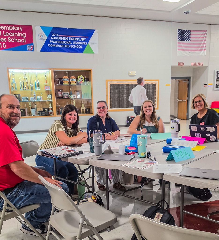 A group of adults are seated at tables in a classroom. The room is brightly lit, with several banners hanging on the wall.