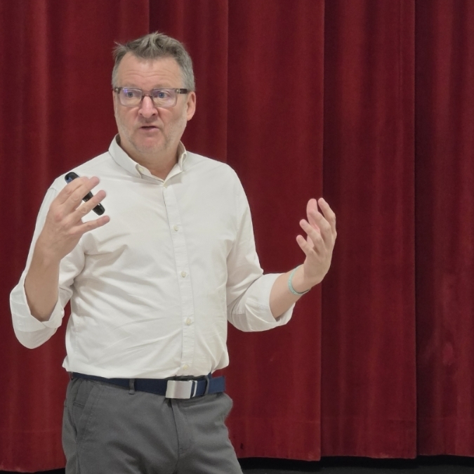 A man in a white shirt and gray pants, wearing glasses, speaks and gestures with a pen.