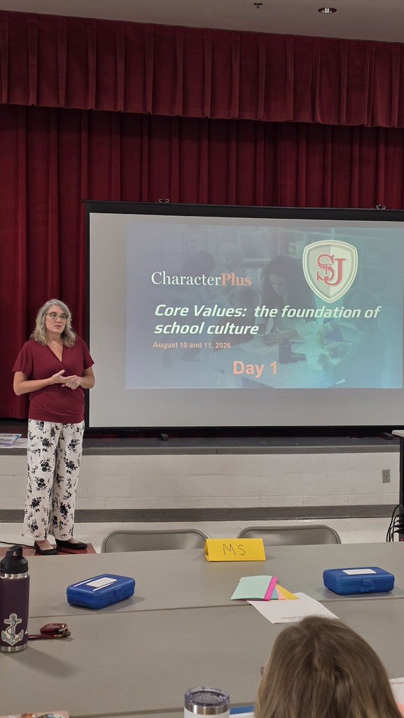 A woman presents in front of a projector screen showing a logo. Behind her is a red curtain.