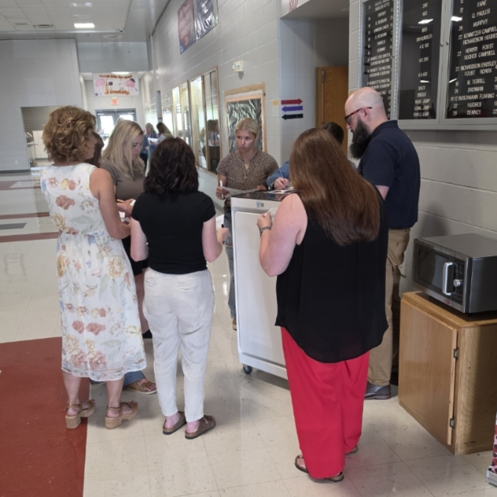 A group of people stands in a school hallway, interacting with a staff member at a counter.
