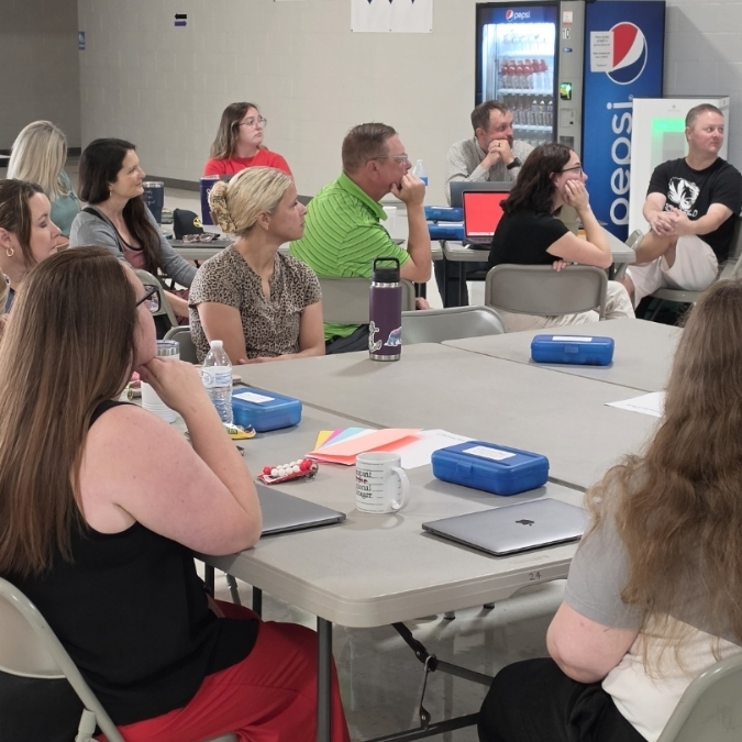 Group of people sitting at tables with laptops, water bottles, and a vending machine in the background.