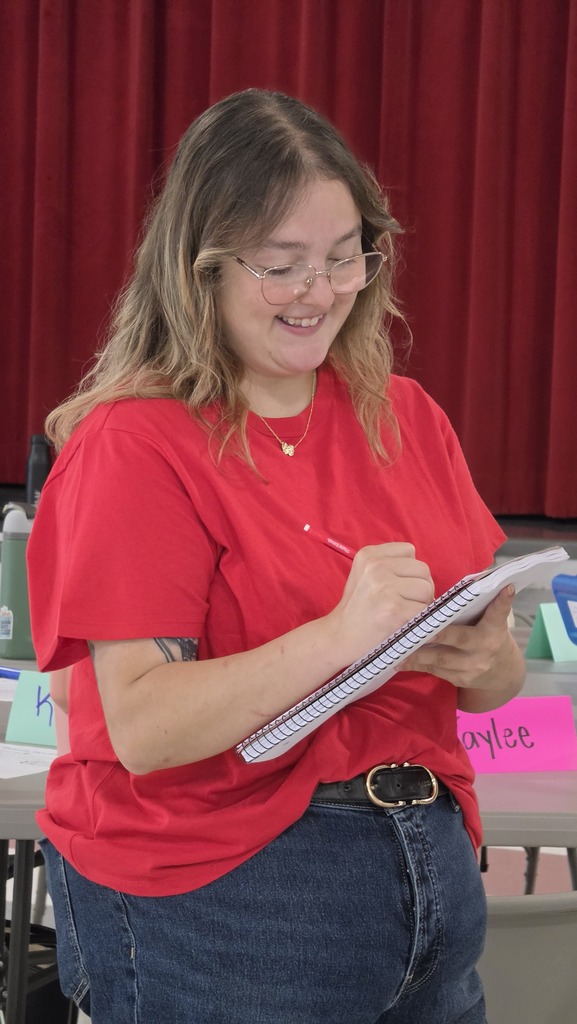 Woman in a red shirt and glasses holds a notebook, writing and smiling in front of a red curtain.