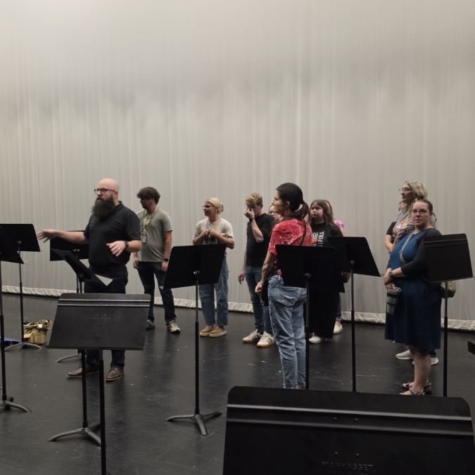 A group of people in a rehearsal room with musical instruments and a white backdrop.