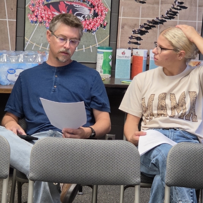 Two people seated on chairs, one with glasses holds a paper while the other looks at them. Glass wall with posters in the background.
