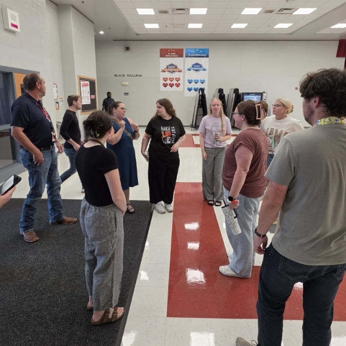 An overhead view of people in a hallway; some stand, others bend. White walls, red floor sections, and ceiling lights.