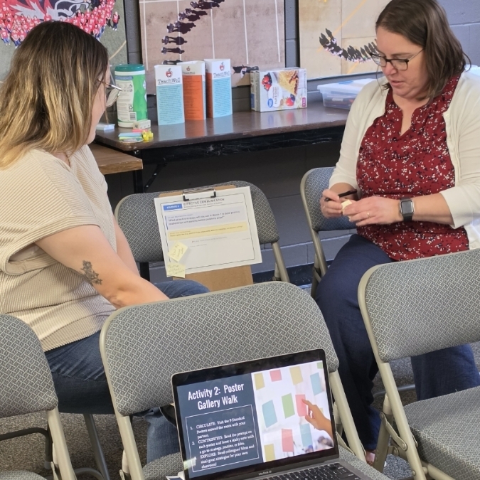 Two women sit in chairs, one with a laptop, the other with a phone. Behind them, a table holds items.