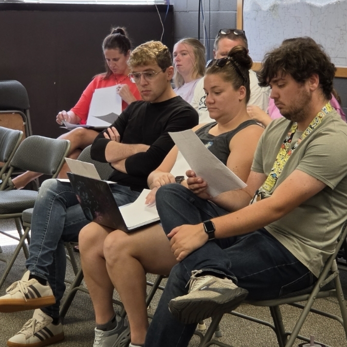 Several people are seated in chairs, facing forward. A person holds papers and another wears a watch.