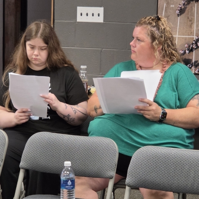 Two women, one with dark hair and a black top, and the other with blonde hair and a green shirt, sit next to each other. Both hold papers.